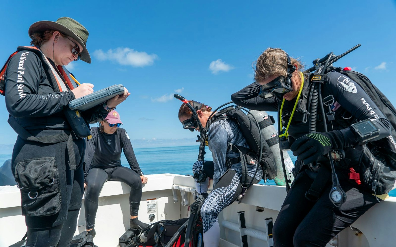 A group of people on a boat, three of them wear scuba gear and one holds a clipboard