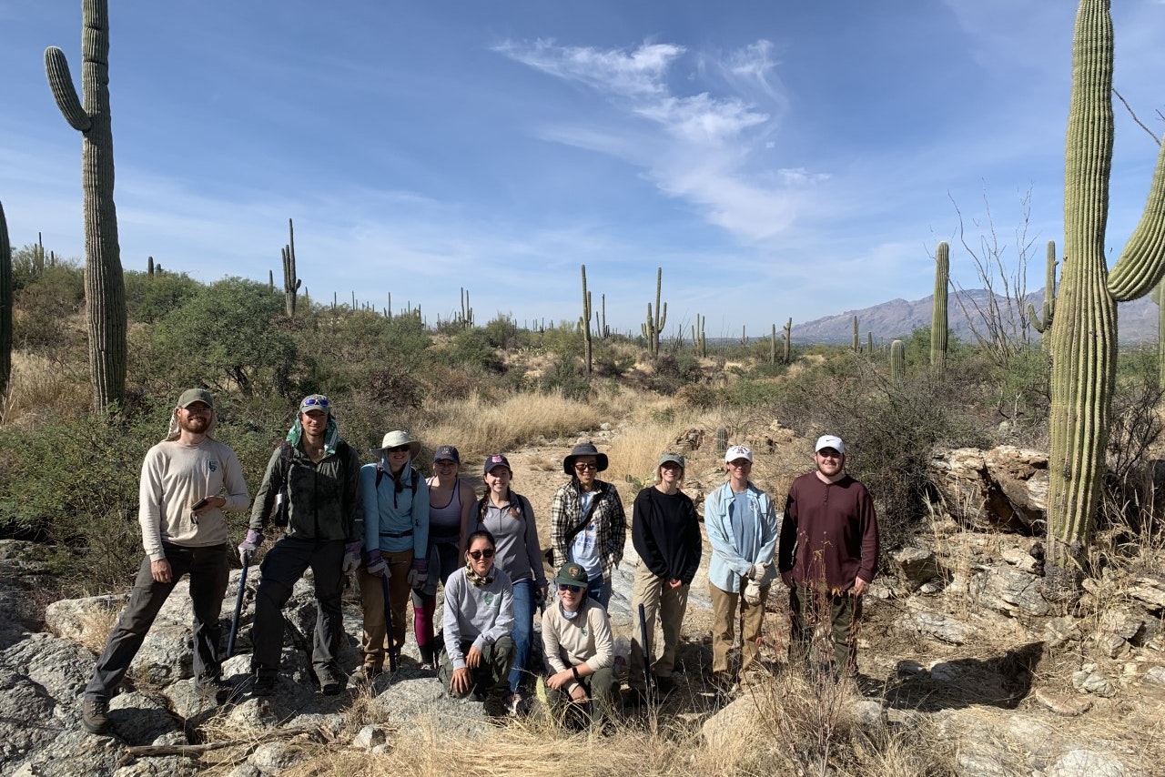 A group of people pose for a group photo in a desert landscape, framed by Saguaro cacti