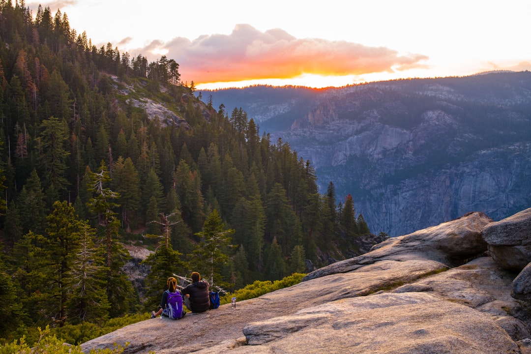 Two people sit on a large rock and overlook a forest at sunset