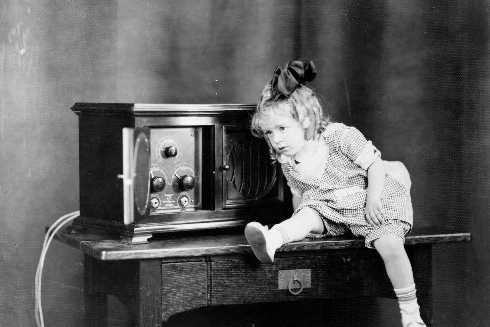 A historic photograph of a child sitting on a tabletop, leaning towards a radio