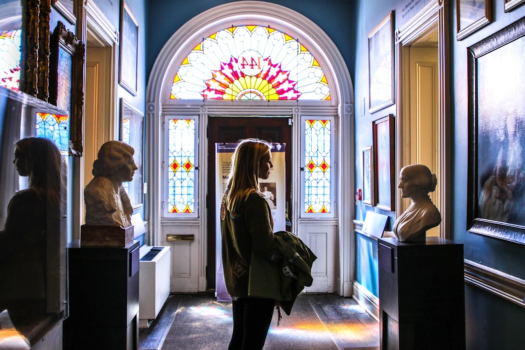 Visitor stops in a hallway to look at a painting, back-lit by sun coming through stained glass windows.