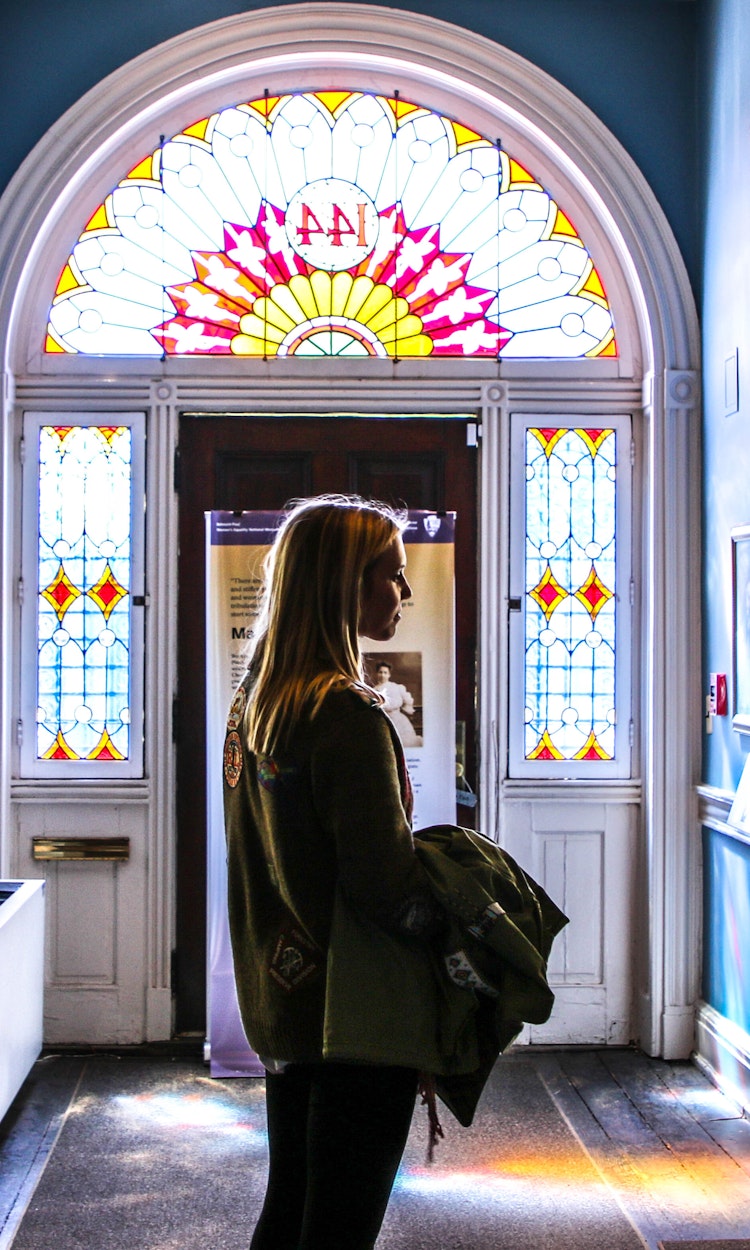 Visitor stops in a hallway to look at a painting, back-lit by sun coming through stained glass windows.