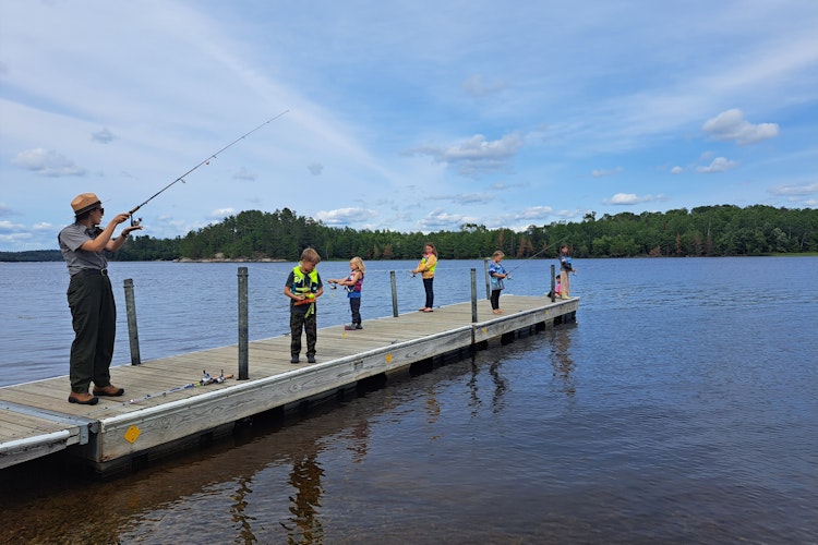 A park ranger, in uniform, stands on a dock with a handful of students as they all fish