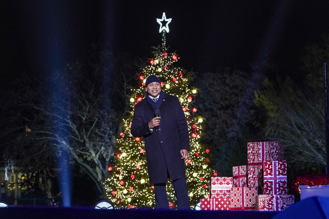 LL COOL J stands in front of an illuminated tree and presents wrapped in red paper. He holds a microphone and smiles.