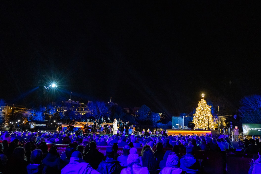 A wide shot of the National Christmas Tree lighting, with the tree illuminated and a gathered crowd watching a speech