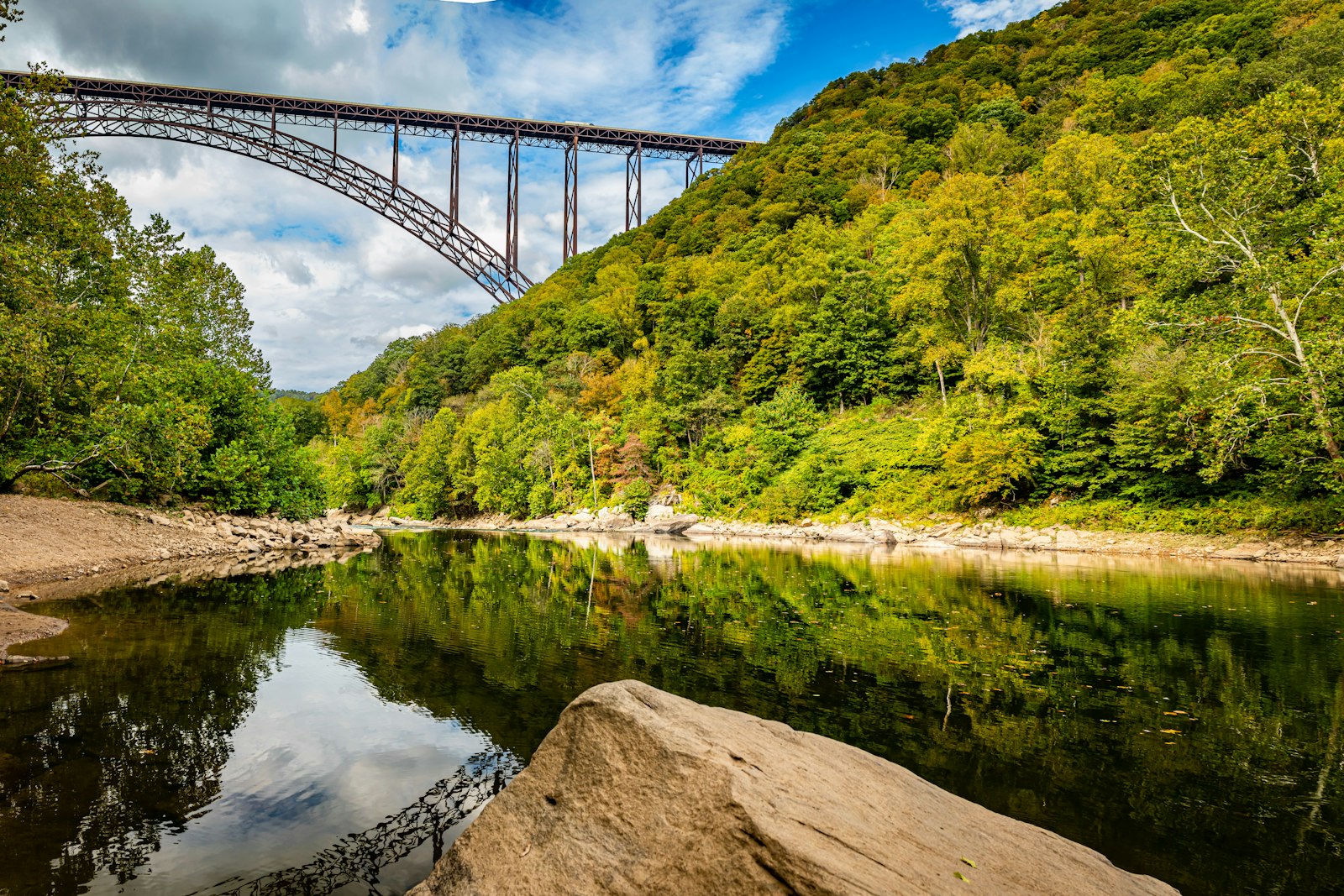 A rock sits in the foreground at the edge of a still river. In the distance, a large elevated steel bridge connects two mountaintops