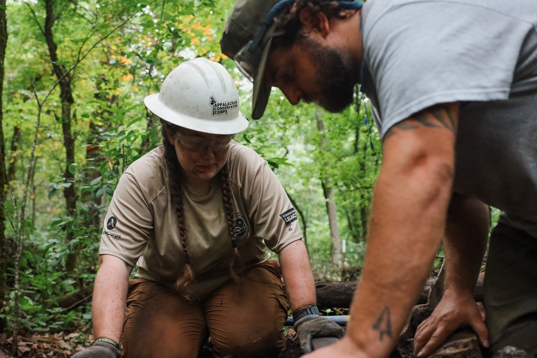 Two people kneel down to work on something. One holds a log. Both wear hats and safety gloves