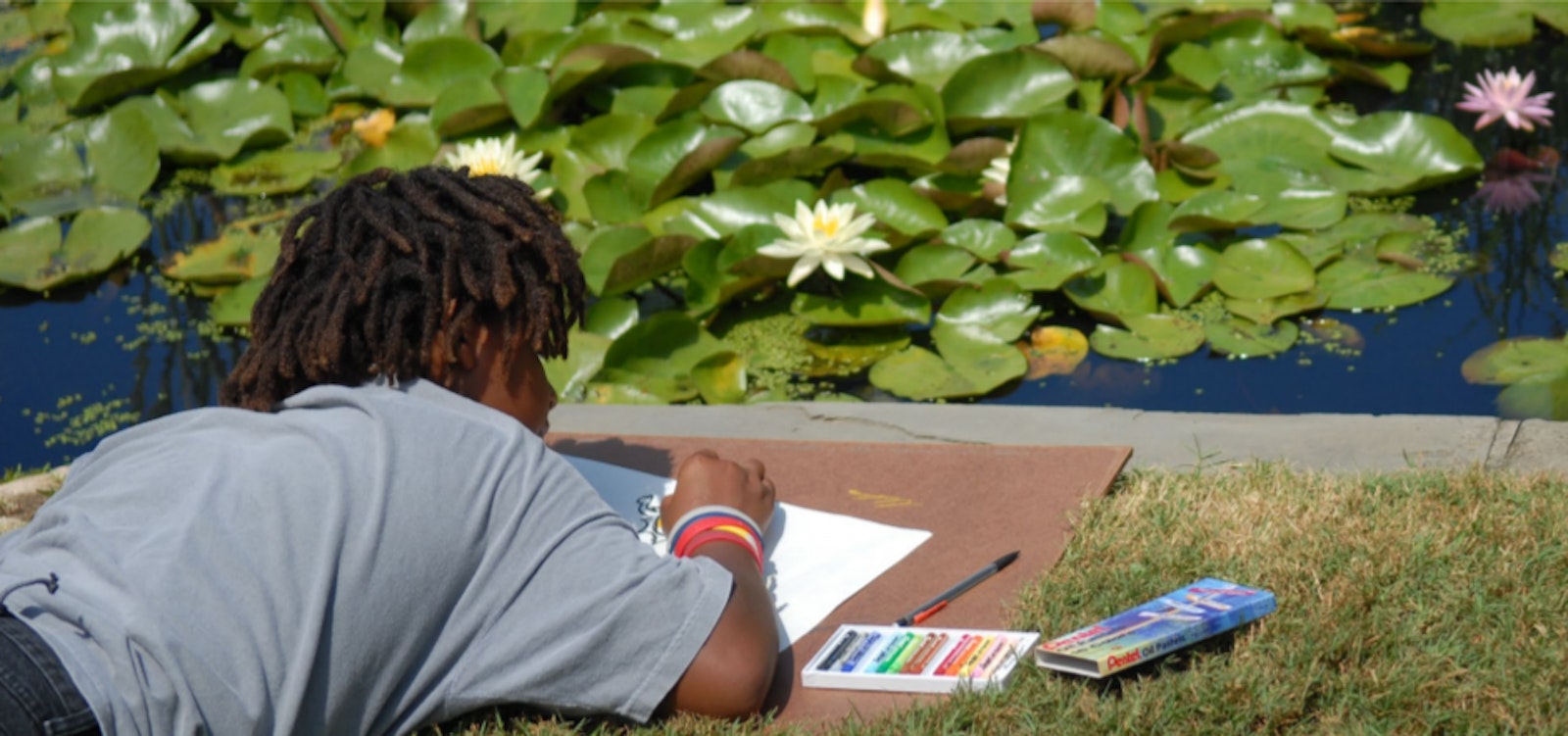 A kid sketches as they lay on their stomach on the edge of a pond