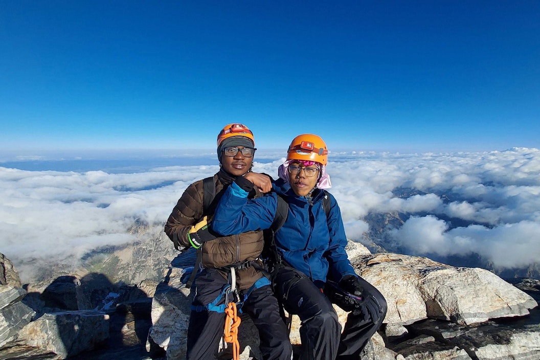 Two people sit at the top of a mountain, above clouds. They wear orange helmets and one curls a bicep