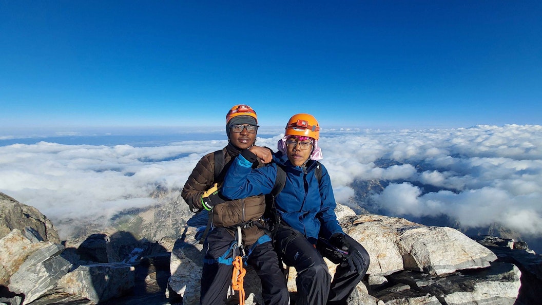 Two people sit at the top of a mountain, above clouds. They wear orange helmets and one curls a bicep
