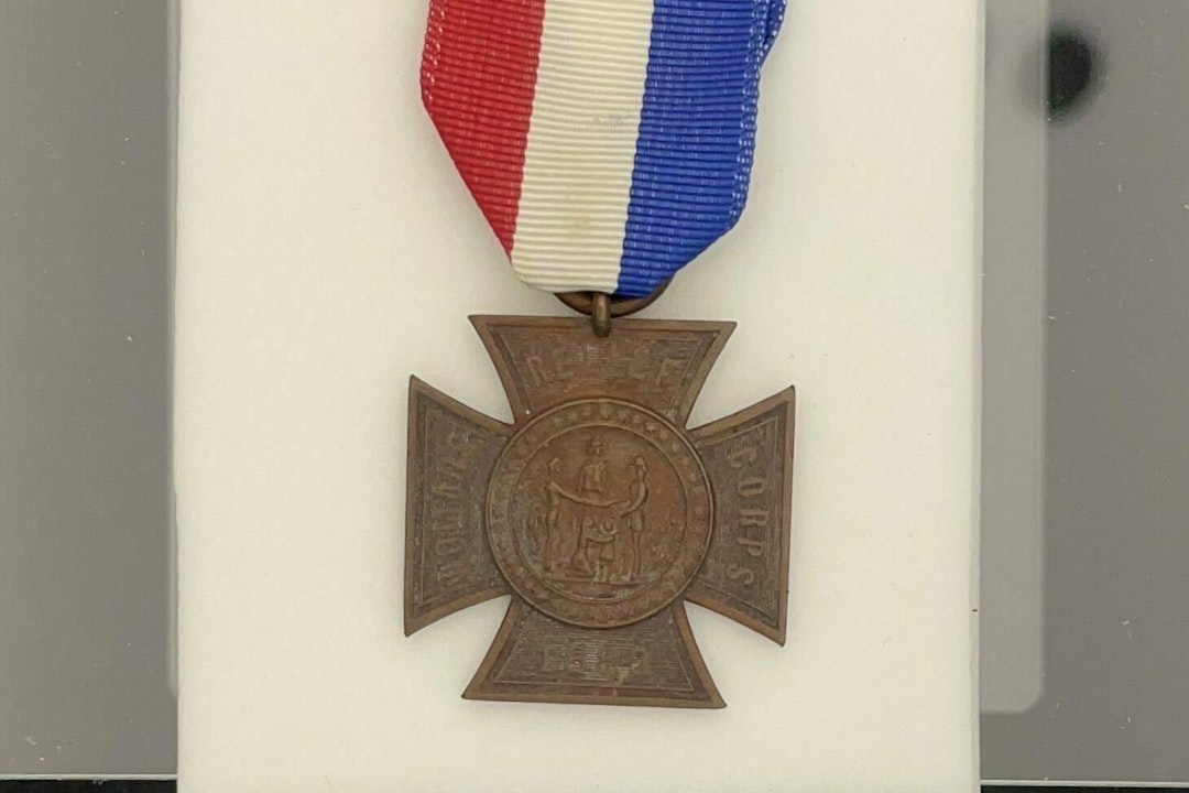 A badge in a box hangs from a red, white, and blue ribbon. At the top, the ribbon is clipped with a pin that says F.C.L.
