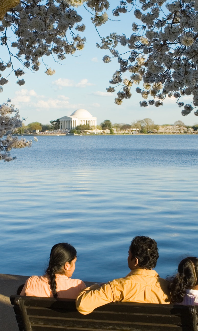 A family sits on a bench at the edge of water. Across the water, a stone, domed memorial can be seen