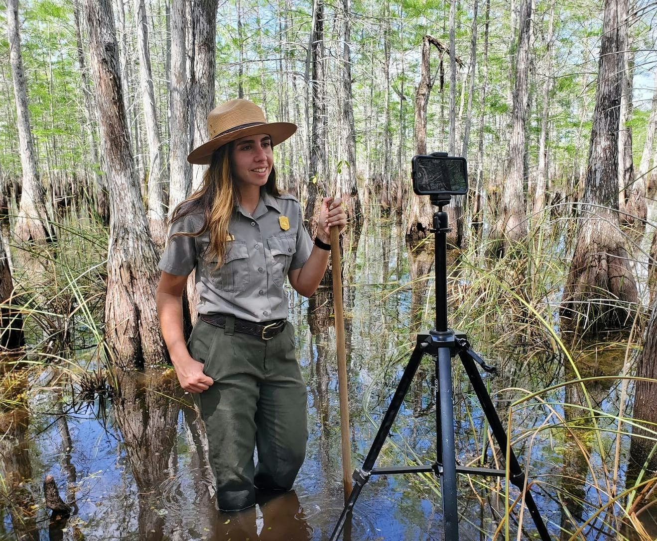 Ranger, in uniform, waves and smiles at a camera on a tripod. She stands in a cypress swamp