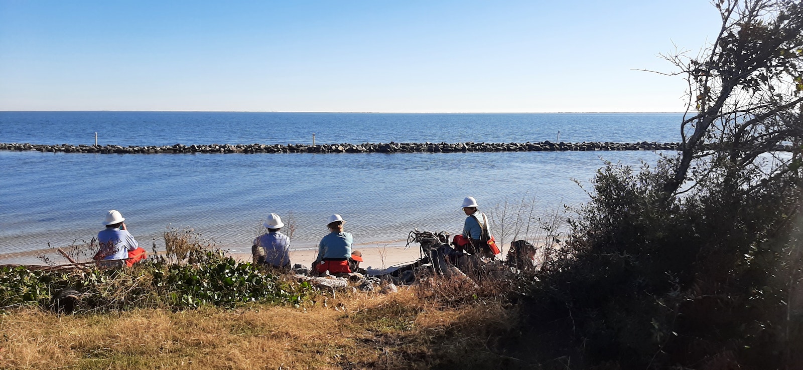 A group of people sit on a shoreline, wearing hardhats