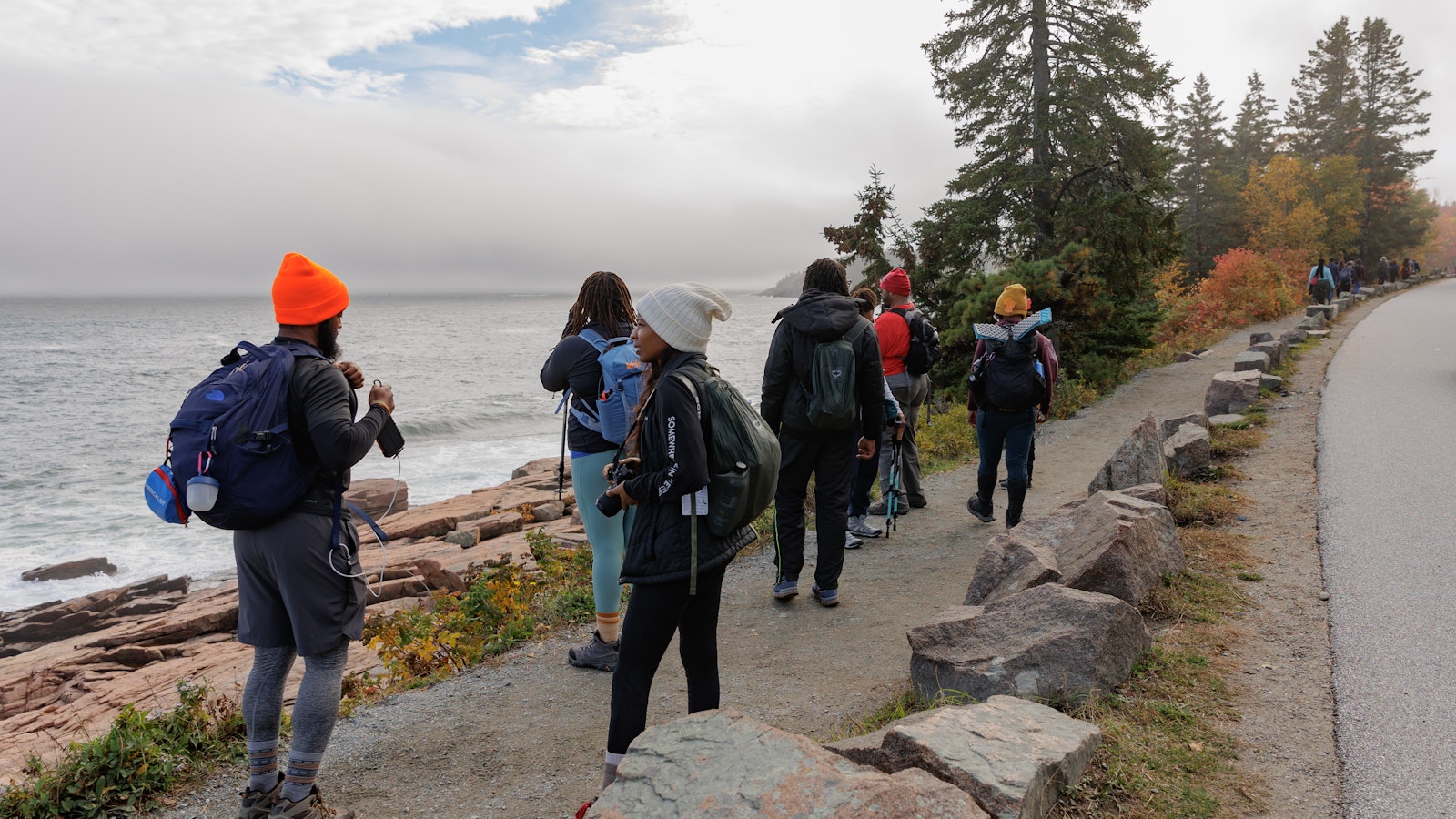 A group of people, wearing hiking gear, walk along a stone path that traces a shore