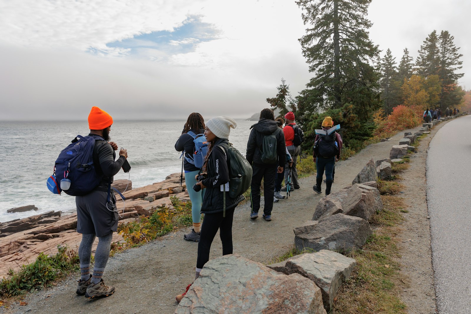A group of people, wearing hiking gear, walk along a stone path that traces a shore