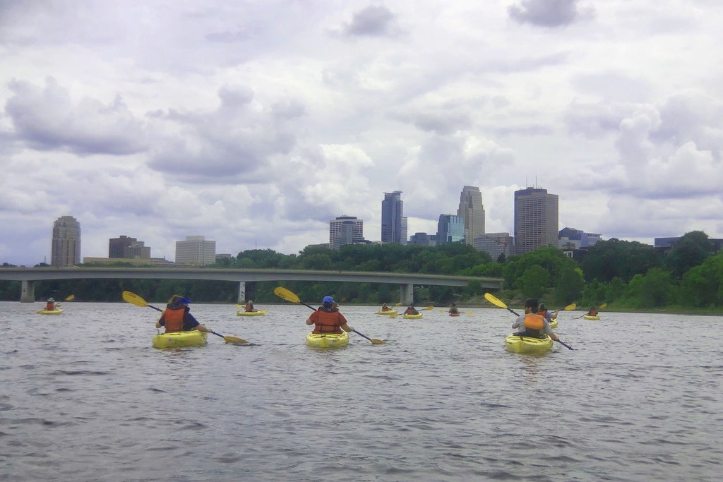 A group of people kayak on a river towards a metropolitan skyline