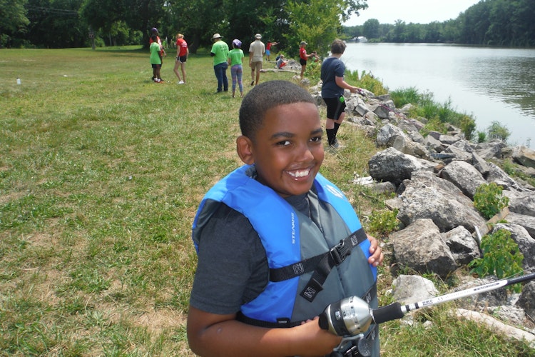 A young visitor smiles at the camera, he holds a fishing rod and wears a life vest
