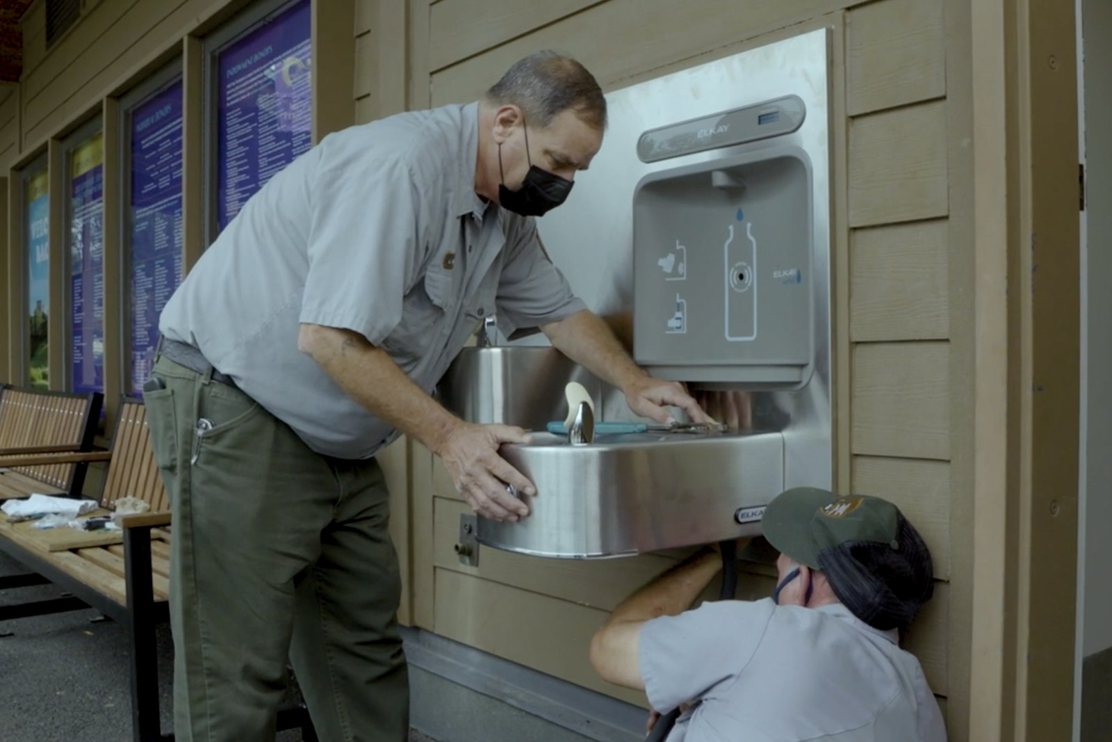 Two uniformed rangers hold and install a water bottle refill station on the exterior of a building, next to a row of benches