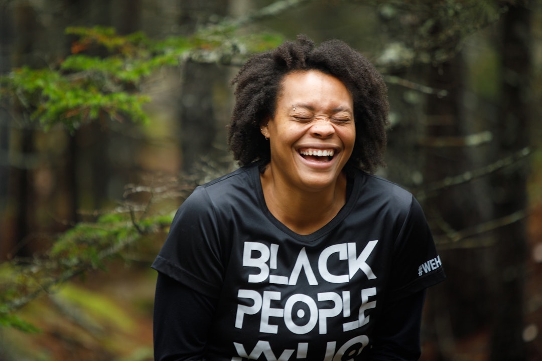 A person smiles, shutting their eyes and crinkling their nose. Their t-shirt reads "Black People Who..."