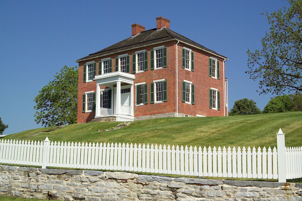 Two story red brick house sits on a hill of freshly mown grass, surrounded by a white picket fence