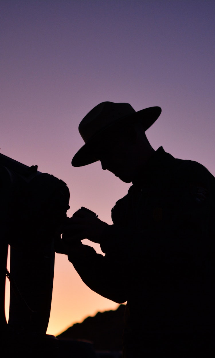 Against a sunset, a ranger in uniform tends to a telescope