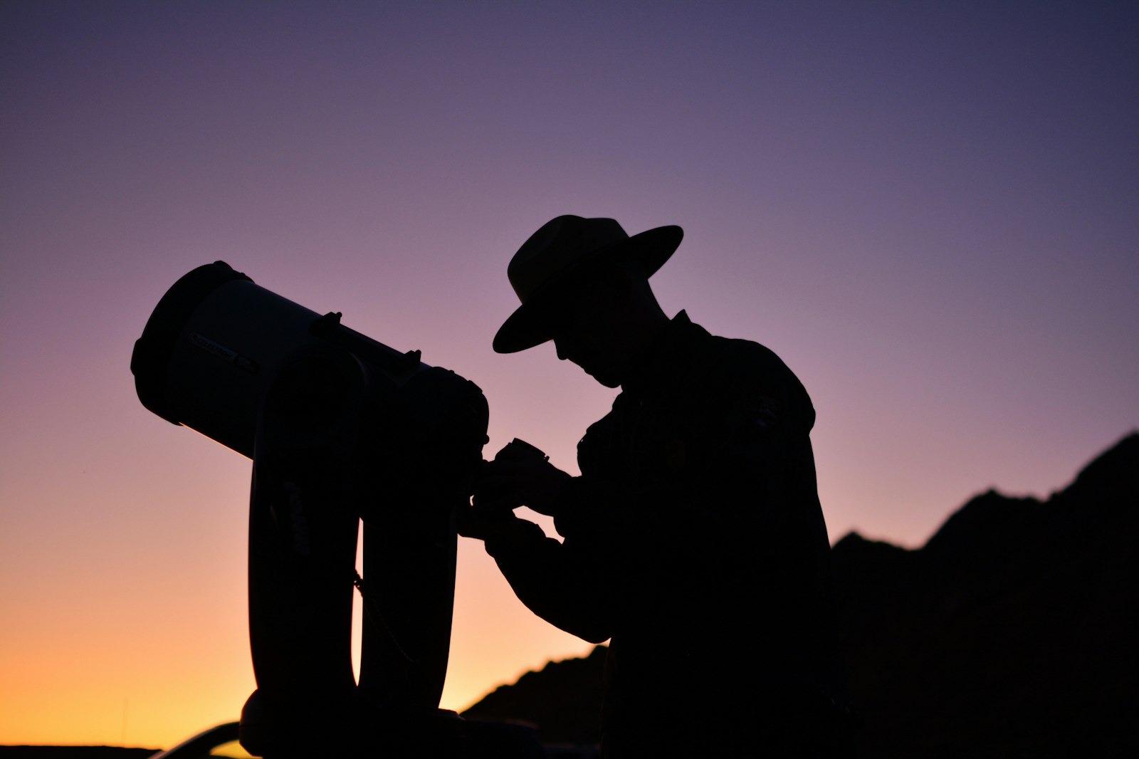 Against a sunset, a ranger in uniform tends to a telescope