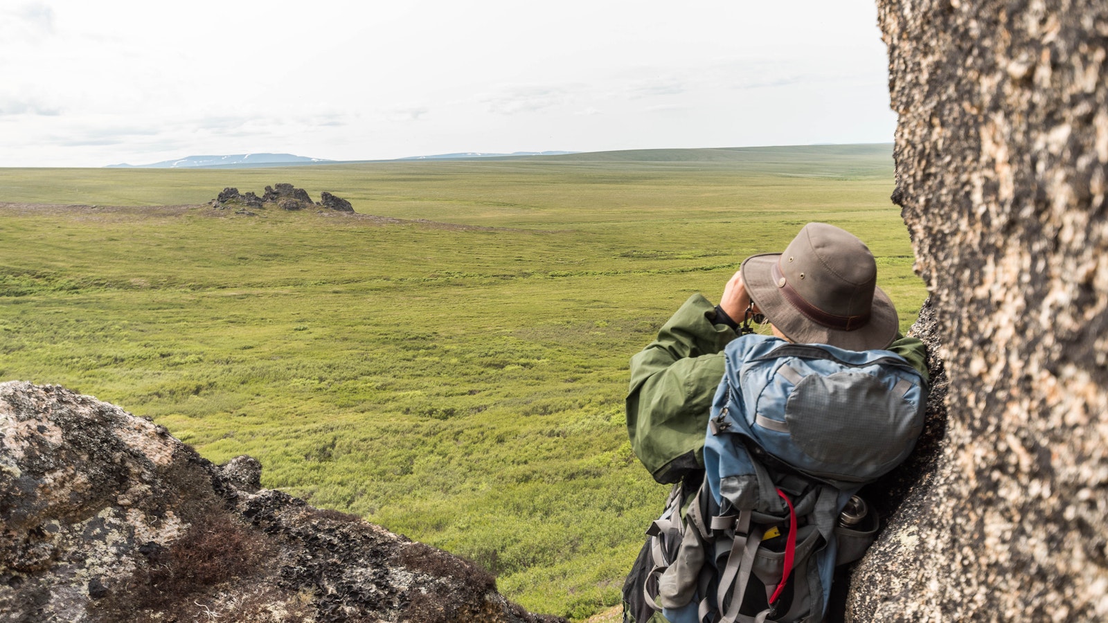 From behind, a female hiker leans against a tor and uses binoculars to look across a vast green landscape.