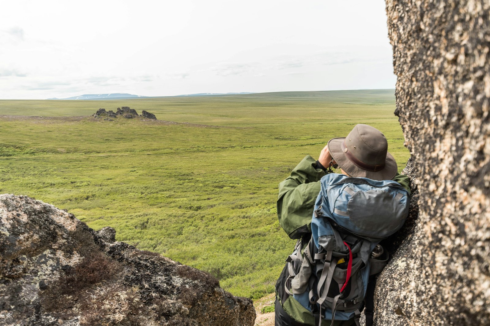 From behind, a female hiker leans against a tor and uses binoculars to look across a vast green landscape.