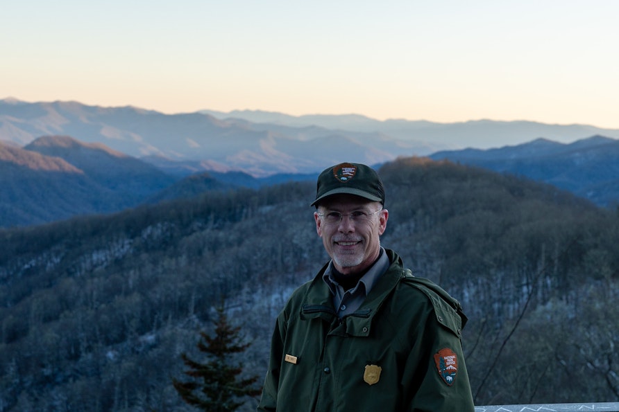 Bill Stiver stands, in his park ranger uniform, in front of a beautiful snowy landscape