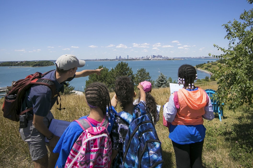 A group on an island point to the Boston skyline across the harbor