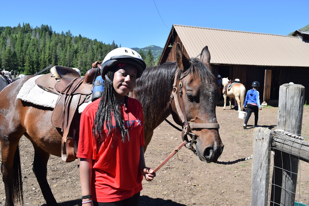 A person, wearing a helmet, holds onto the reins of a horse at a horse ranch