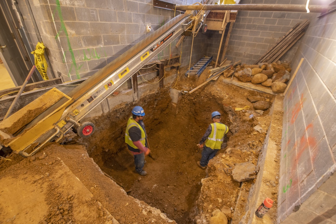 An interior construction pit, with two people wearing high-visibility vests, gloves, and hard hats standing in the pit