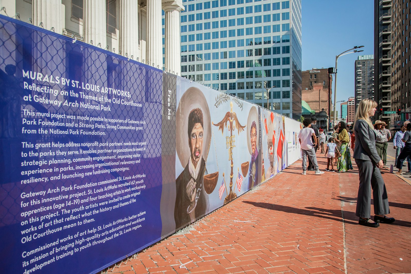 People stand and talk on a red brick sidewalk. On a fence surrounding an old courthouse is a color-printed screen with art and a long blurb of text