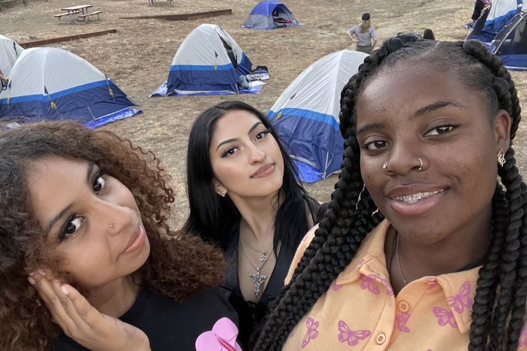 Three young women take a selfie. In the background are tents