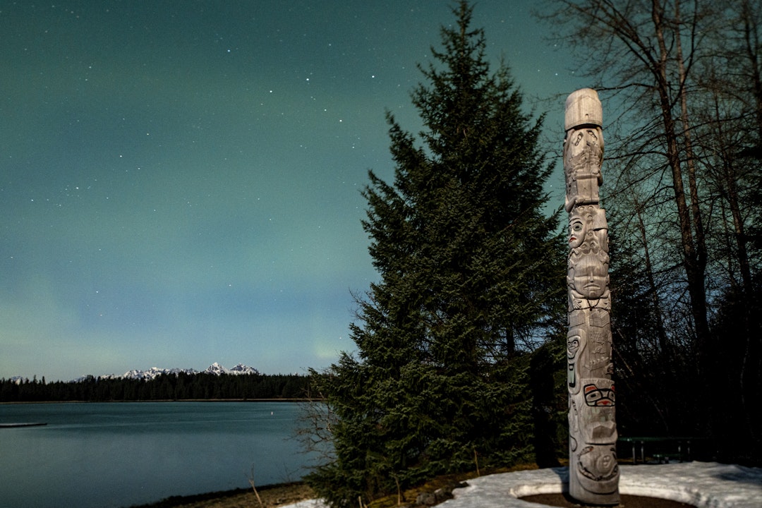 Against the backdrop of the northern lights and pine trees stands a tall cedar totem pole