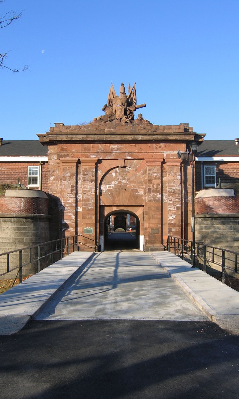 View of a bridge leading to the entrance of a brownstone fort gatehouse with and bald eagle sculpture atop it.
