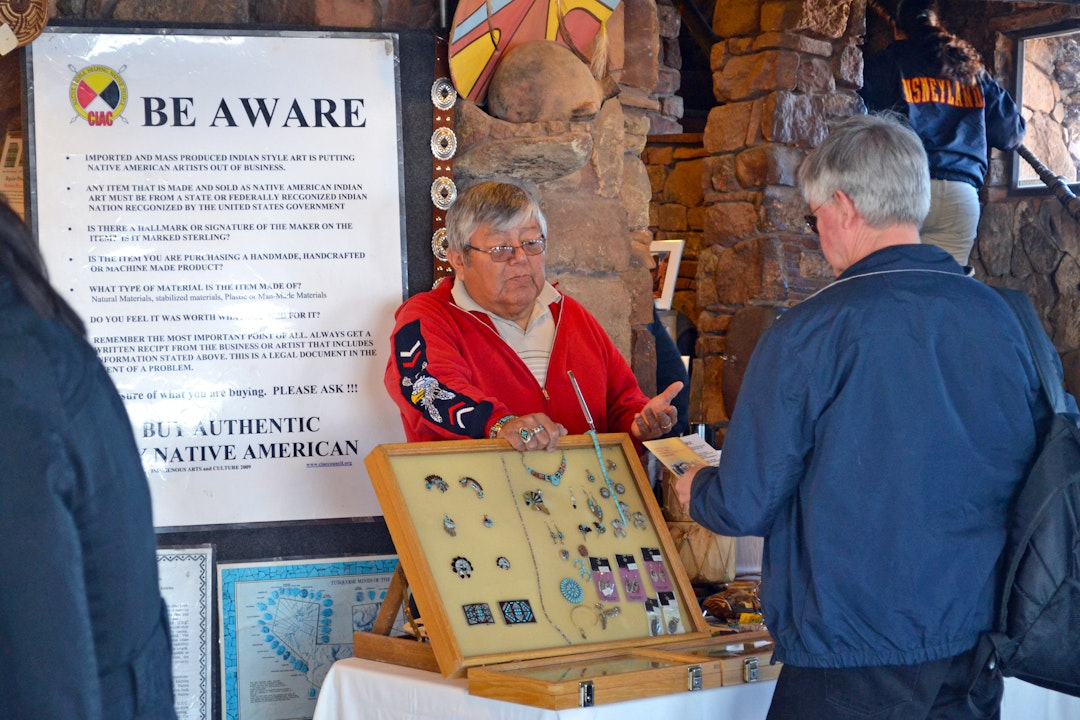 A person holds up a board, showcasing an array of jewelry, and speaks to someone standing in front of them