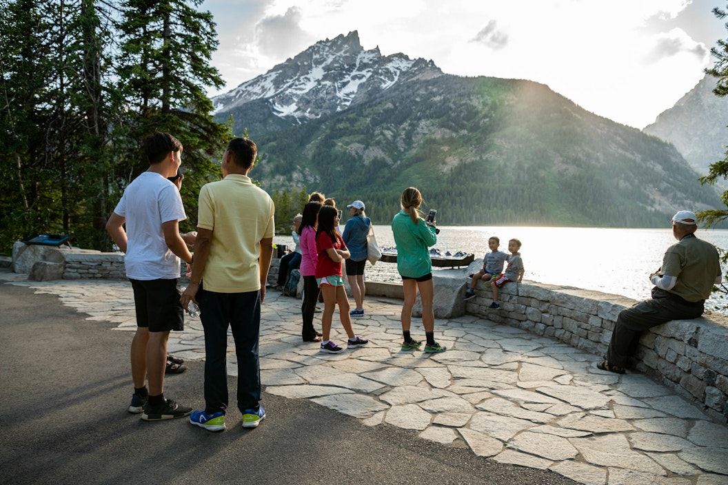 A group of people stand and sit along a low stone bench overlooking a still lake surrounded by snow-capped mountains