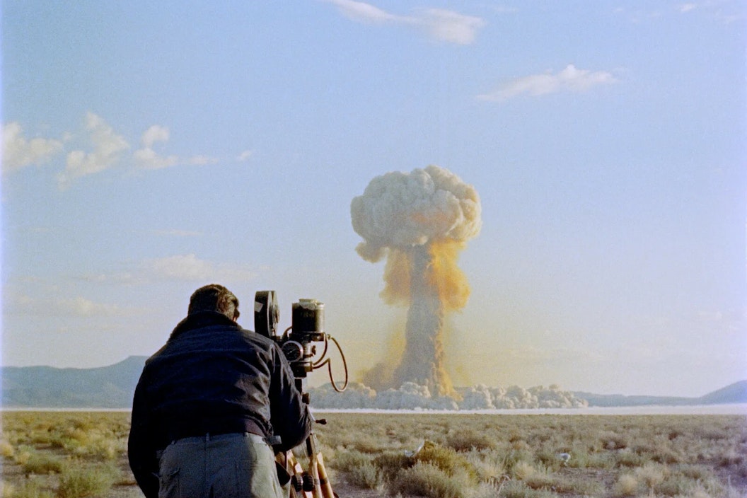 A man stands at an old film camera and films a nuclear explosion in the distance