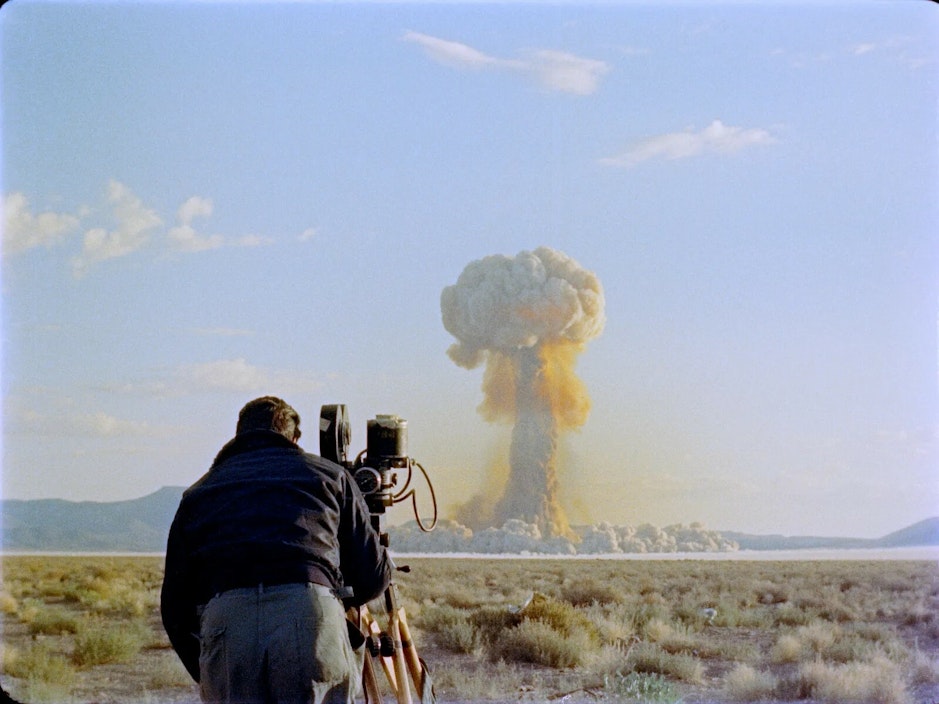 A man stands at an old film camera and films a nuclear explosion in the distance
