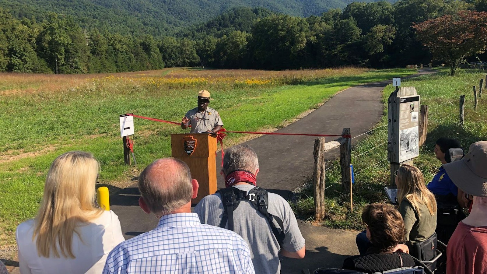 A group of people gather to listen to a person speak from behind a lectern, in front of a paved trail