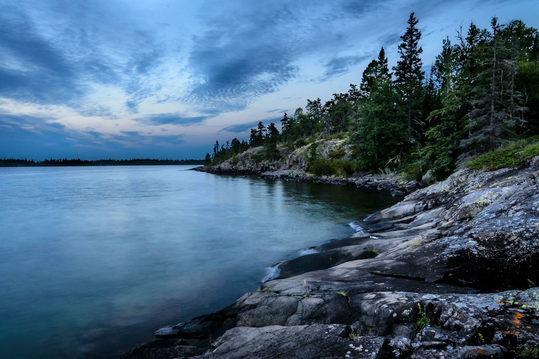 A calm lake just off the shore of a rocky shoreline, dotted with evergreen trees