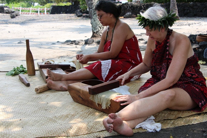 Two women in traditional clothing beat kapa while sitting on a luahala mat.