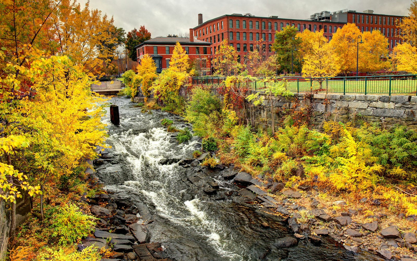 A small waterfall and stream rush along rocks, surrounded by orange, red, and yellow foliage. In the distance is a large red-brick building