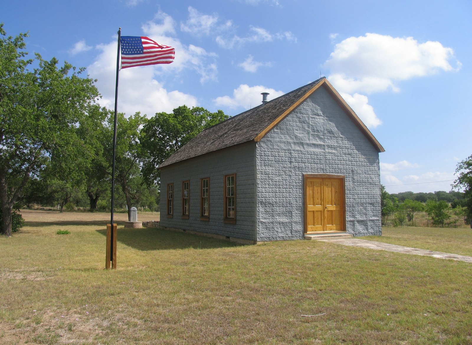 One-room schoolhouse with grey-blue facade and four windows on the side. In the lawn in front of the school flies an American flag on a flagpole
