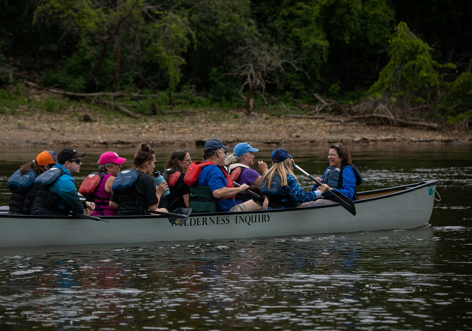 A group of people sit in a canoe, on a river, and smile