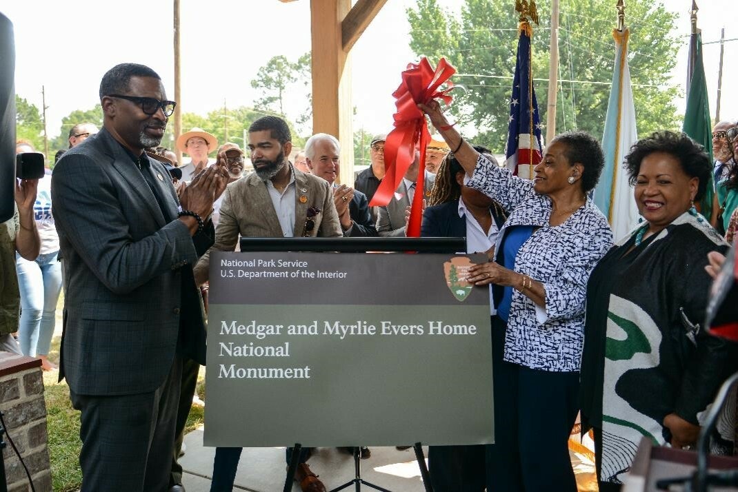 A group of people stand around a sign, smiling. One woman lifts a red ribbon off the sign. The sign reads: Medgar & Myrlie Evers Home National Monument