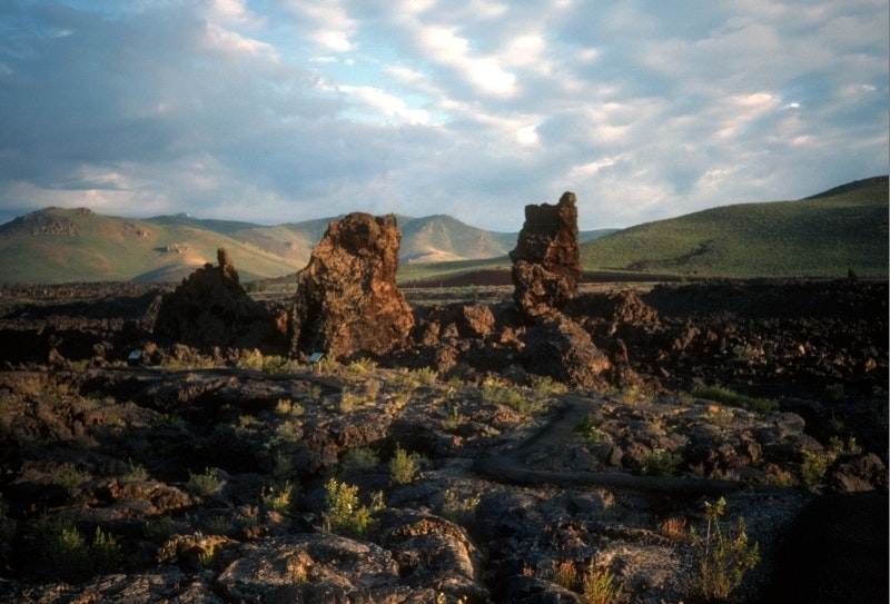 three tall chunks of reddish black rocks between a chunky black lava field and a smooth vegetated lava flow with green mountains in the distance