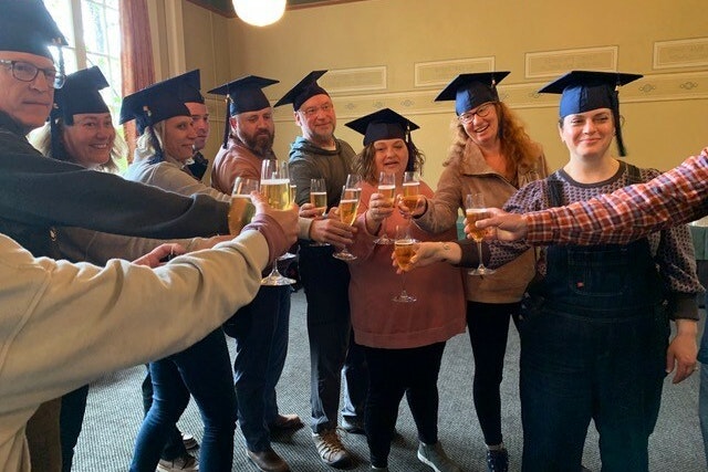 A group of people, wearing graduation hats and standing in a circle, raise glasses and cheers